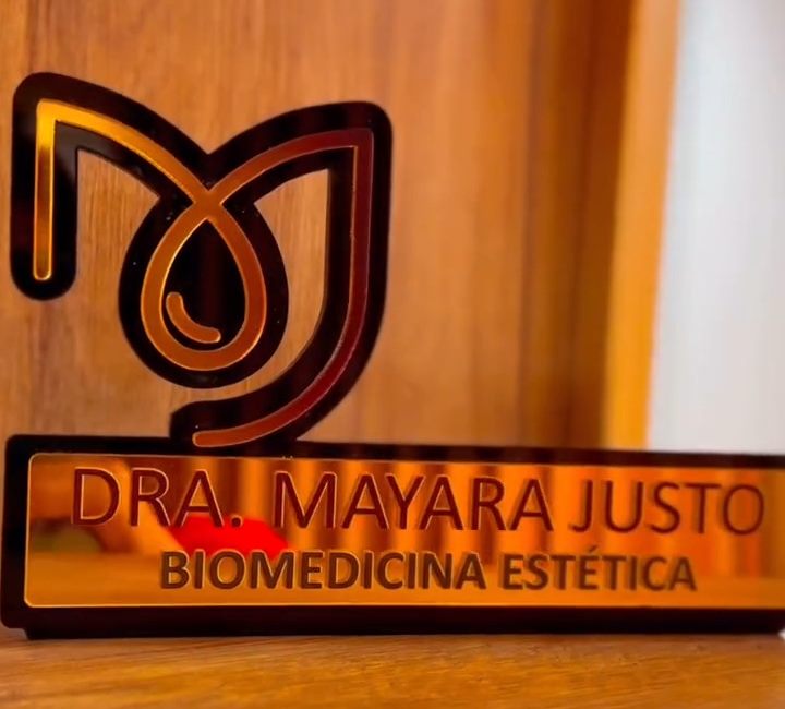 Placa dourada com o monograma e o nome Dra. Mayara Justo, Biomedicina Estética